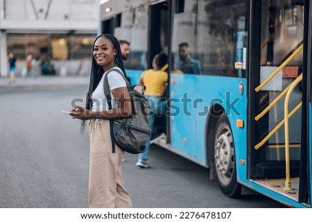 Portrait of an african american woman looking at the camera while waiting at a bus stop. Young black female using public transportation while going to school or work. Using smartphone for bus schedule Royalty-Free Stock Photo #2276478107