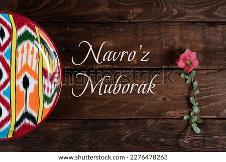 Rishtan Uzbek national plate and   pink spring flower with congratulatory text on wooden background. Postcard for Navruz