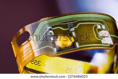 Closeup of electronic flexible printed circuit on plastic strip curled to ring on purple background. Green and yellow lines and small electric components on flex PCB strip from disassembled earphones. Royalty-Free Stock Photo #2276486023