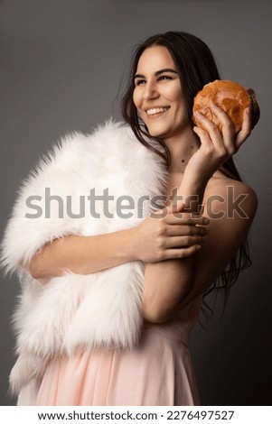 Studio portrait of a beautiful caucasian woman wearing a pink gown and a faux white fur shawl. She is holding a cheeseburger and posing. She has brown eyes and long brown hair.