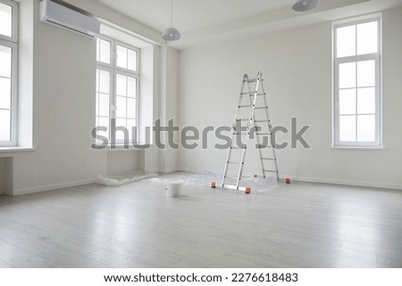 Modern apartment after renovations. New empty office interior or living room at home with white freshly painted walls, step ladder, paint bucket, plastic on the floor, big windows, and air conditioner
