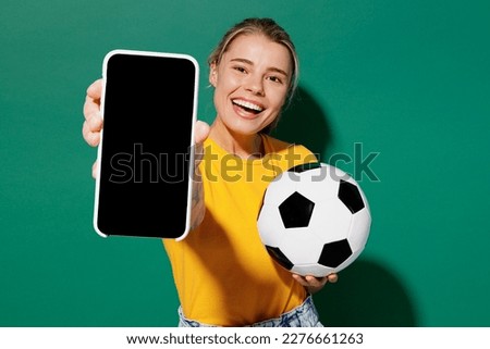 Young woman fan wear basic yellow t-shirt cheer up support football sport team hold soccer ball watch tv live stream use mobile cell phone closeup blank screen isolated on dark green background studio
