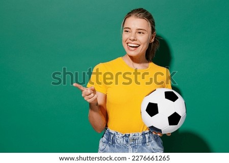 Young woman fan wear basic yellow t-shirt cheer up support football sport team hold in hand soccer ball watch tv live stream point index finger aside on area isolated on dark green background studio Royalty-Free Stock Photo #2276661265