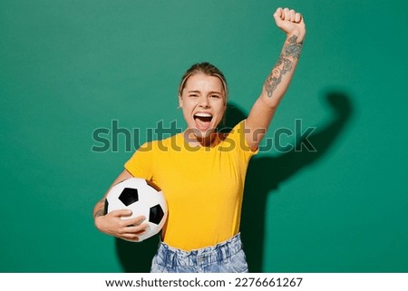 Young excited woman fan wear basic yellow t-shirt cheer up support football sport team hold in hand soccer ball watch tv live stream do winner gesture shout isolated on dark green background studio Royalty-Free Stock Photo #2276661267