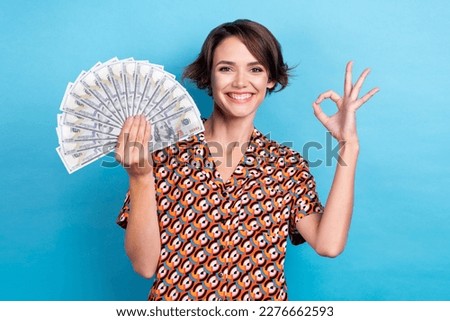 Photo of charming confident woman wear print blouse showing okey sign holding money fan isolated blue color background