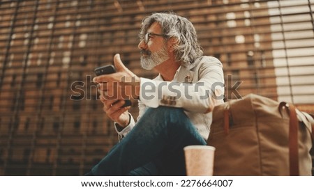 Mature businessman with beard in eyeglasses wearing gray jacket is using cell phone. Middle aged manager scrolling information on his smartphone while sitting outside the office Royalty-Free Stock Photo #2276664007