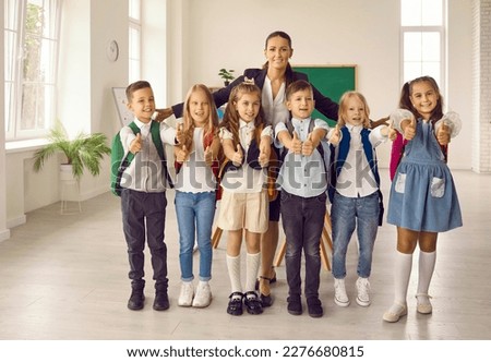 Happy female enthusiastic teacher hugs students of her elementary class. Group photo for parents. Positive kids gesturing thumbs up with both hands all together. Education concept. Royalty-Free Stock Photo #2276680815