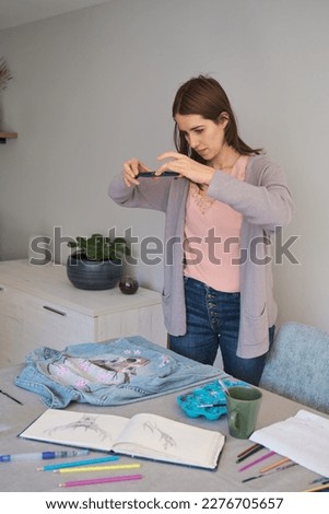 Caucasian woman taking a photo of her art design in a denim jacket. Unique clothing designs.