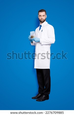 Full size fullbody portrait of attractive stylish scientist in white lab coat, black pants, shoes, tie holding test tubes with multi-colored liquid, looking at camera, isolated on grey background Royalty-Free Stock Photo #2276709885