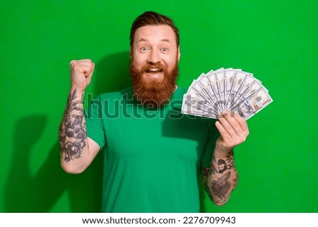 Photo of satisfied funny guy ginger beard dressed stylish t-shirt hold dollars raising fist win bet isolated on green color background