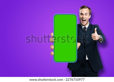 Excited cheerful business man in black suit, show cell phone, mobile smartphone. Happy businessman hold cellphone with green chroma key screen isolated violet purple background. Thumb up. Great app ad