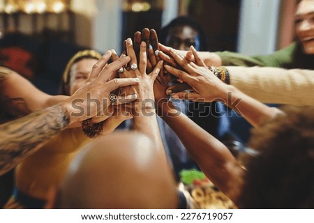 A diverse group of friends, in their forties, with various ethnicities and tattoos, gather around a table for dinner. They enthusiastically join hands in a high-five, blurred faces and focus on hands