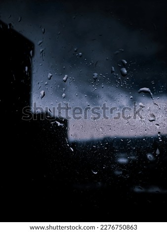 A rainy day environment, raindrops on the colorful window through the defocused cloudy black sky and blurry city buildings, rainy day nature view from the window at almost night