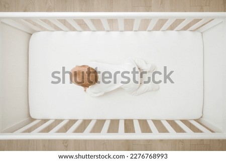 Baby in white bodysuit try crawling on knee and arms on mattress in wooden crib at home room. Top view. 5 to 6 months old infant development. Royalty-Free Stock Photo #2276768993