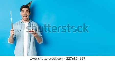 Happy young man celebrating birthday in party hat, holding b-day cake and smiling, standing over blue background.