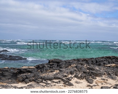 a coastal landscape on Fuerteventura, very windy and very high waves, turquoise water and white sand with lava rocks in the water, with sunshine and few clouds