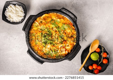 Delicious Brazilian fish moqueca with tomato, onion, olive oil, coriander and orocum seed. Made in clay pot on white table and black background. Top view.