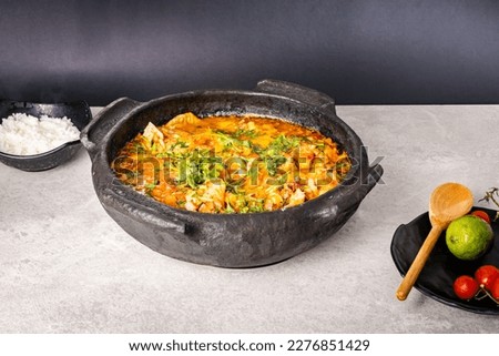 Delicious Brazilian fish moqueca with tomato, onion, olive oil, coriander and orocum seed. Made in clay pot on white table and black background. With rice. Royalty-Free Stock Photo #2276851429