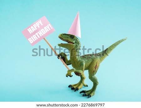 CUte dinosaur in birthday hat holding Happy Birthday sign on blue background. CUte birthday greeting card idea concept.
