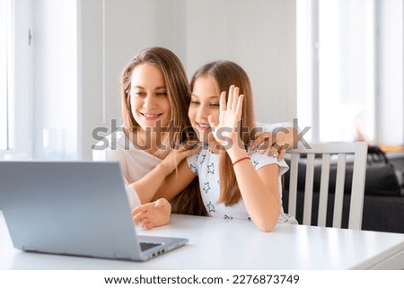 Mom and daughter having online video cal with school teacher - they looking to the laptop monitor and waving hand. Distant education concept.