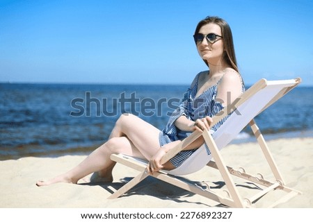Happy young brunette woman relaxing on a wooden deck chair at the ocean beach while smiling and wearing fashion sunglasses. The enjoying vacation concept.