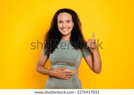 Happy brazilian young woman in sportswear, keeps one hand on her stomach, the other one shows thumb up gesture, demonstrating that she feels good, looks at camera, smiles, isolated yellow background Royalty-Free Stock Photo #2276917031
