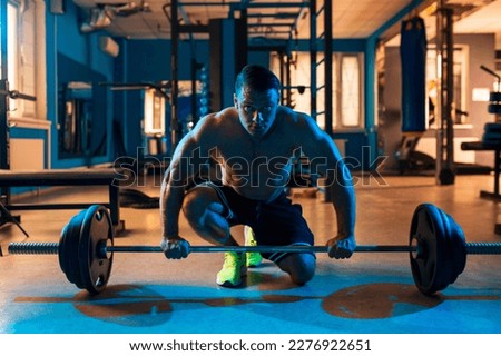 Athletic fit sporty muscular caucasian man on colored blue light sitting on his knee and holding barbell with weight disc plates in his hands topless in black shorts in contemporary gym