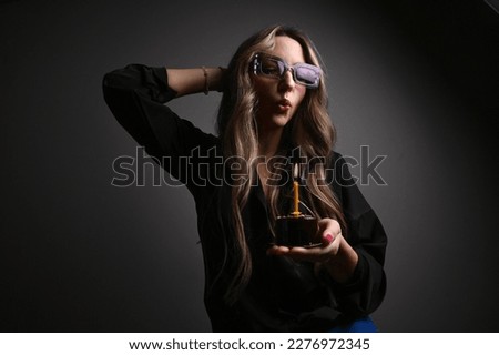 Portrait of stylish woman in purple glasses holding birthday cake against dark grey wall. Woman blowing out candle on cake in hands
