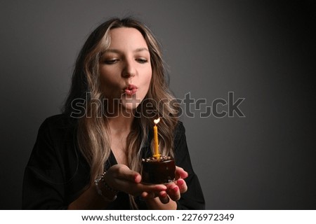 Portrait of woman holding birthday cake against dark grey wall. Woman blowing out candle on cake in hands. Make a wish.