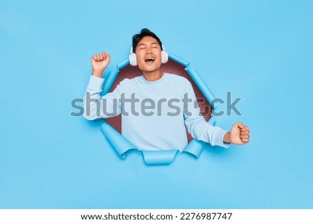 Happy young guy in white sweatshirt and headphones standing inside blue studio background, student listening music and singing cheerfully, youth lifestyle concept, copy space