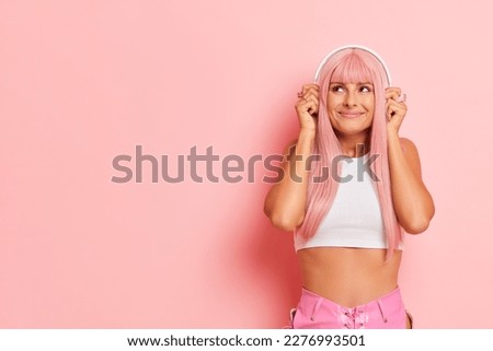 Happy smiling young woman in white top holding both hands up on her earphones, pretty model listening nice music posing on studio pink copy space, relax concept, high quality photo