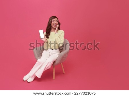 Happy young Asian woman sitting on sofa feeling happiness and gesture showing mobile smartphone blank screen on isolated pink background.