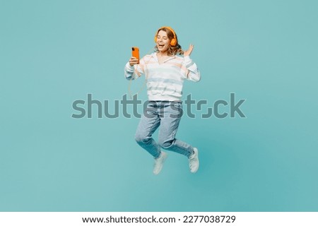 Full body side view young woman wears striped hoody headphones listen to music use mobile cell phone jump high isolated on plain pastel light blue cyan background studio portrait. Lifestyle concept