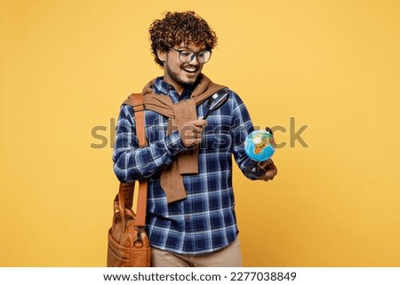 Young smiling teen Indian boy IT student he wear casual clothes glasses bag hold globe Earth map use magnifying glass isolated on plain yellow color background High school university college concept