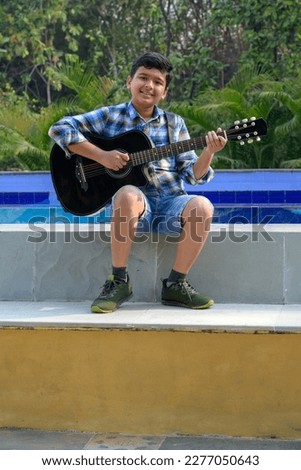 Indian asian boy playing acaustic guitar while sitting beside swimming pool