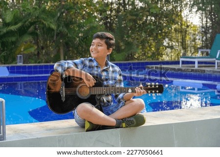 Indian asian boy playing acaustic guitar while sitting beside swimming pool