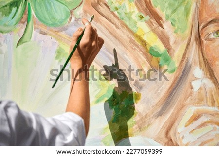 Girl artist hand holds paintbrush and draws green nature landscape on canvas at outdoor art painting festival, paintings art picture process. Woman artist paints atmospheric surreal picture