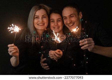 Happy family in black - woman, man and little girl, with a sparkler in hand the New Year's Christmas at home. The girl at the father on hands. New year 2023.