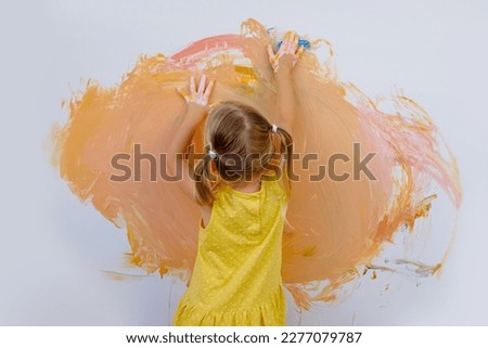 small child, blonde girl 3 years in yellow dress paints with her hands orange paint on white wall, childish naive drawing, gouache, acrylic, happiness childhood, creative development Royalty-Free Stock Photo #2277079787