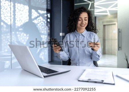 Cheerful and successful business woman at workplace inside office, using phone and bank credit card to buy online in online store, Hispanic woman working at work with laptop.