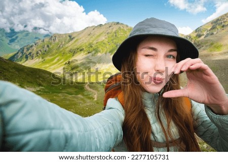 Cute happy girl making heart shaped hand gesture looking at camera smiling funny, caucasian young single woman blogger laughing face showing love sign symbol, mountains, nature, closeup portrait