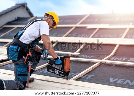 The carpenter nails a timber board using an electric nailer while working on a roof. Royalty-Free Stock Photo #2277109377