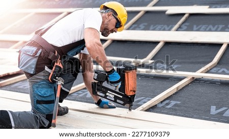 The carpenter nails a timber board using an electric nailer while working on a roof. Royalty-Free Stock Photo #2277109379