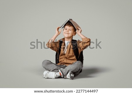 Glad little european boy with backpack puts book on head, sits on floor, enjoy learning isolated on gray studio background. Schoolboy emotions, fun, study and education, ad and offer Royalty-Free Stock Photo #2277144497