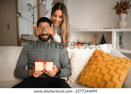 Smiling international millennial couple celebrating birthday, anniversary, holiday, wife closes eyes to husband and gives gift box in living room interior. Surprise, relationship and love at home