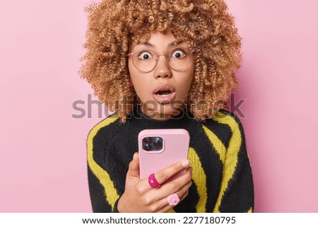 Shocked speechless man with curly hair holds mobile phone stares with omg expression wears transparent eyeglasses and casual jumper reacts to something impressive isolated over pink background Royalty-Free Stock Photo #2277180795