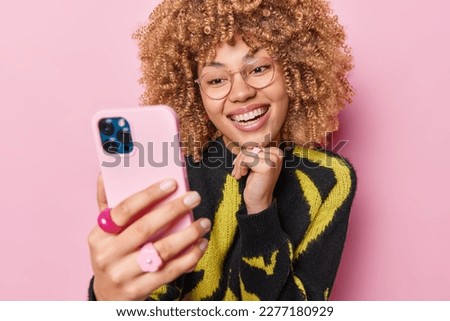 Image of young positive woman with curly hair takes picture on smartphone talks on video call app keeps hand under chin dressed in black and yellow jumper isolated over pink studio background