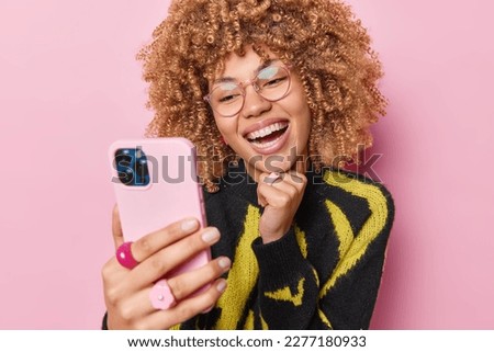Photo of young cheerful adult woman with curly hair smiles broadly makes photo on smartphone or has funny conversation online wears transparent glasses and jumper isolated over pink background
