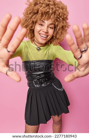 Vertical shot of cheerful woman with curly hair keeps hands towards camera smiles gladfully wears black corset and skirt has happy mood isolated over pink background. Positive emotions concept