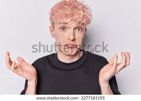 Hesitant pink haired guy shrugs shoulders feels uncertain looks confusingly at camera purses lips dressed in casual black t shirt doesnt know right answer isolated over white background. So what Royalty-Free Stock Photo #2277181193
